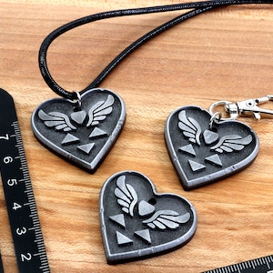 Puede incluir: Tres colgantes de coraz&oacute;n plateados con un s&iacute;mbolo alado y un dise&ntilde;o triangular. Un colgante est&aacute; en un collar de cord&oacute;n negro, y los otros dos est&aacute;n en llaveros.