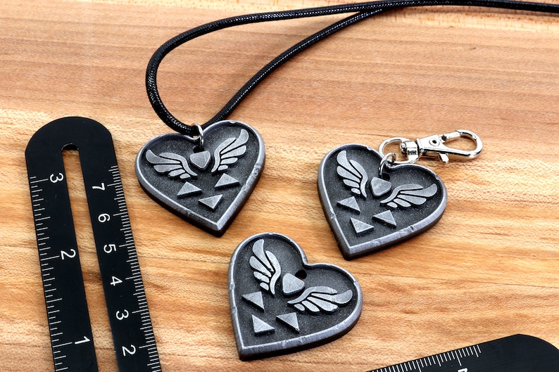 Puede incluir: Tres colgantes de coraz&oacute;n plateados con un s&iacute;mbolo alado y un dise&ntilde;o triangular. Un colgante est&aacute; en un collar de cord&oacute;n negro, y los otros dos est&aacute;n en llaveros.