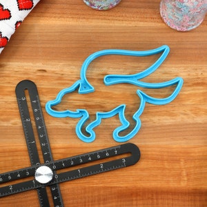 May include: A blue plastic cookie cutter shaped like a winged fox. The cutter is on a wooden surface with a black ruler.
