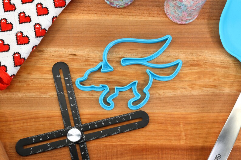 May include: A blue plastic cookie cutter shaped like a winged fox. The cutter is on a wooden surface with a black ruler.