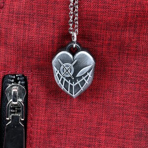May include: A silver heart-shaped pendant necklace with a detailed design of a face with a cross-eye and a smile. The necklace is on a red fabric background.