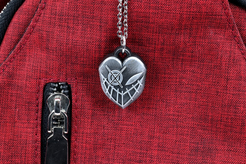May include: A silver heart-shaped pendant necklace with a detailed design of a face with a cross-eye and a smile. The necklace is on a red fabric background.
