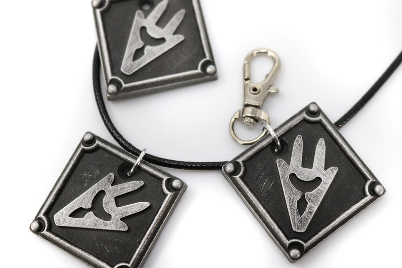 May include: A black necklace with three silver square pendants featuring a stylized silver symbol. The necklace has a silver lobster clasp.