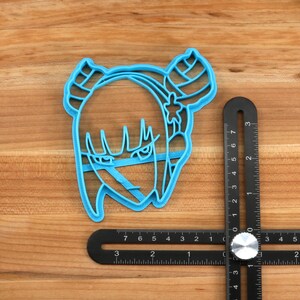 May include: A blue plastic cookie cutter shaped like a cartoon character with horns, a star on its head, and a serious expression. The cutter is 2 inches wide and is sitting on a wooden surface next to a ruler.