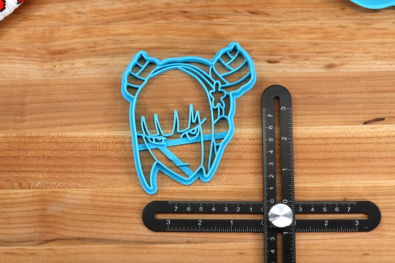 May include: A blue plastic cookie cutter shaped like a cartoon character with horns, a star on its head, and a serious expression. The cutter is 2 inches wide and is sitting on a wooden surface next to a ruler.