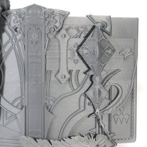 May include: A gray 3D printed model of a book with intricate details and a decorative design. The book has a clasp and a feather bookmark.