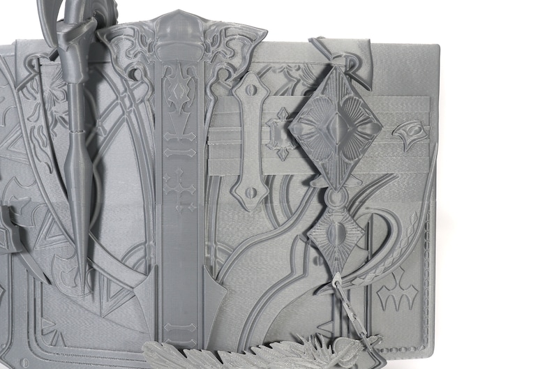 May include: A gray 3D printed model of a book with intricate details and a decorative design. The book has a clasp and a feather bookmark.
