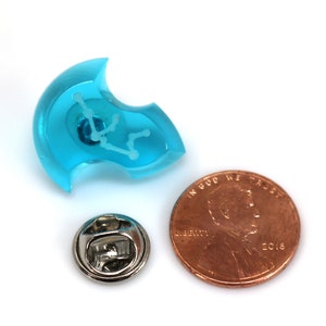 May include: A turquoise blue resin pin with a white outline of the Aquarius constellation. The pin is shown next to a US penny for size comparison.
