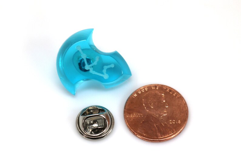 May include: A turquoise blue resin pin with a white outline of the Aquarius constellation. The pin is shown next to a US penny for size comparison.