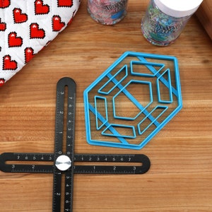 Op de afbeelding: Een blauwe, zeshoekige koekjesuitsteker van plastic met een geometrisch ontwerp. De uitsteker ligt op een houten oppervlak met een zwarte liniaal en een deegroller.