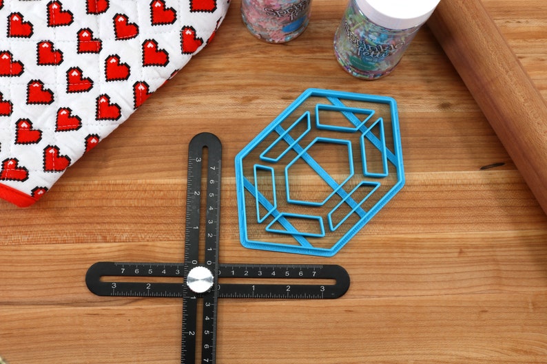 Op de afbeelding: Een blauwe, zeshoekige koekjesuitsteker van plastic met een geometrisch ontwerp. De uitsteker ligt op een houten oppervlak met een zwarte liniaal en een deegroller.