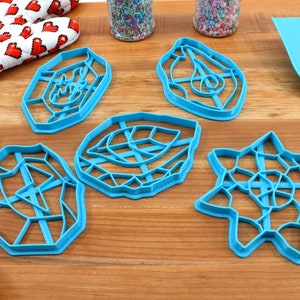 May include: Five blue plastic cookie cutters in different geometric shapes, including a star, a circle, and a leaf. The cutters are on a wooden surface.