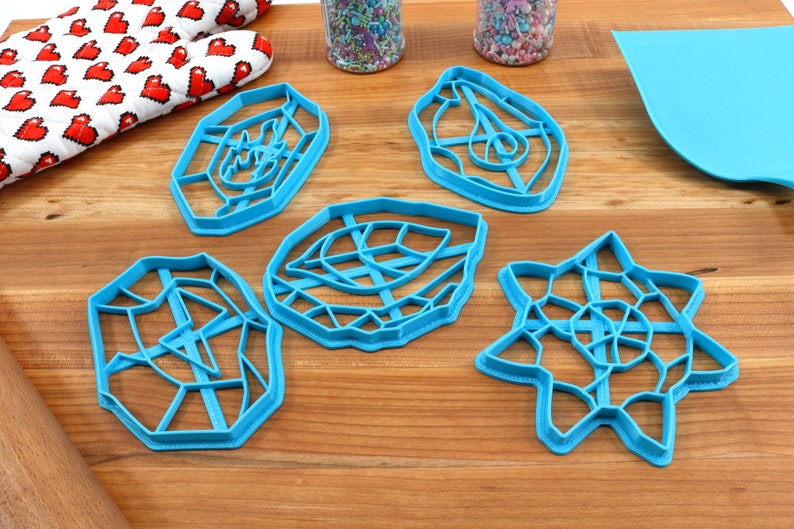 May include: Five blue plastic cookie cutters in different geometric shapes, including a star, a circle, and a leaf. The cutters are on a wooden surface.