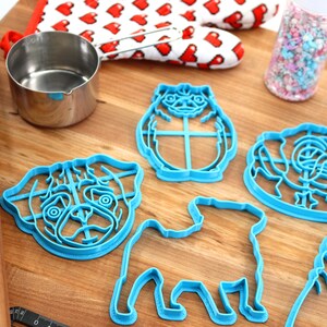 May include: A set of five blue plastic pug-shaped cookie cutters on a wooden surface. The cutters are in various poses, including sitting, standing, and a head-only design.