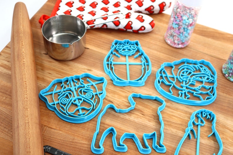 May include: A set of five blue plastic pug-shaped cookie cutters on a wooden surface. The cutters are in various poses, including sitting, standing, and a head-only design.