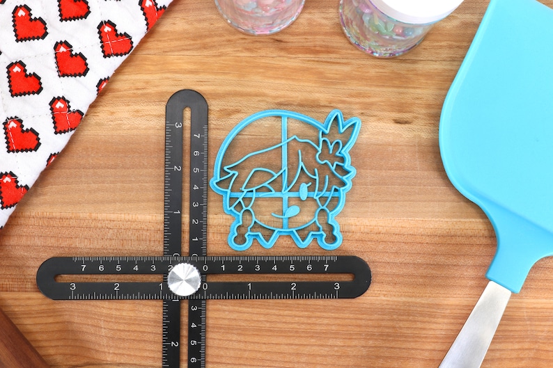 Pu&ograve; includere: Un tagliapasta in plastica blu a forma di personaggio dei cartoni animati con una corona. Il tagliapasta &egrave; su una superficie di legno con un righello e una spatola blu.