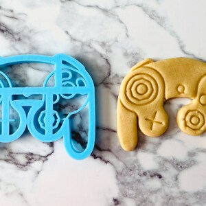 May include: A blue plastic cookie cutter shaped like a video game controller, next to a light brown cookie in the shape of a video game controller.