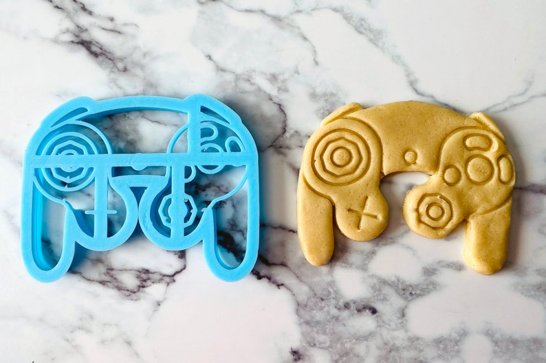 May include: A blue plastic cookie cutter shaped like a video game controller, next to a light brown cookie in the shape of a video game controller.