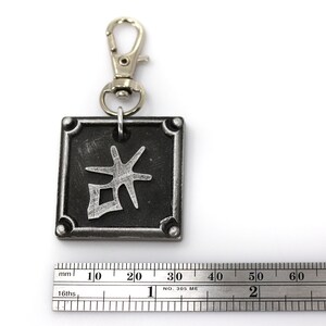 May include: A silver metal keychain with a black square charm featuring a silver kanji character. The charm has a silver border and four silver rivets.