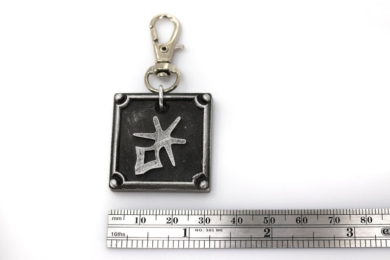 May include: A silver metal keychain with a black square charm featuring a silver kanji character. The charm has a silver border and four silver rivets.