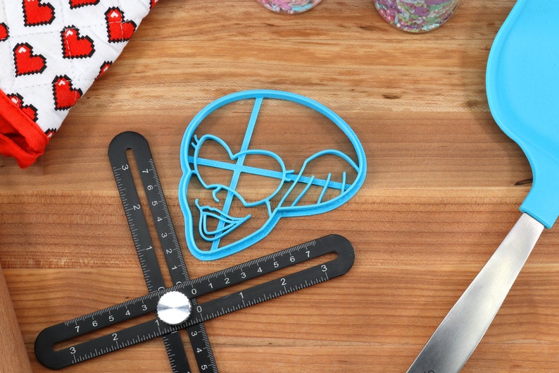 Peut inclure: Un emporte-pi&egrave;ce en plastique bleu en forme de personnage de dessin anim&eacute; avec une t&ecirc;te en forme de c&oelig;ur. Une r&egrave;gle d'angle r&eacute;glable noire et argent&eacute;e est &agrave; c&ocirc;t&eacute; de l'emporte-pi&egrave;ce. Un gant de cuisine &agrave; motifs de c&oelig;urs rouges et blancs est en arri&egrave;re-plan.