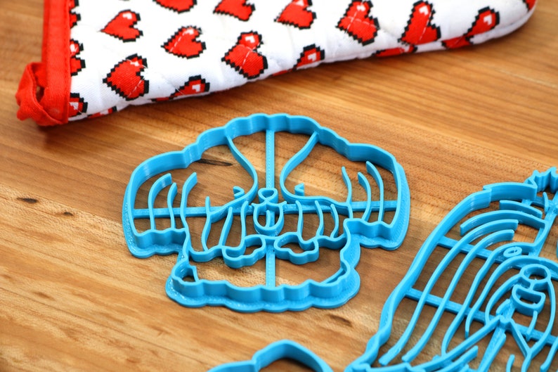 May include: Two blue plastic cookie cutters shaped like a flower and a dog. The cutters are on a wooden surface.