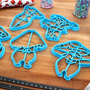 May include: Set of five blue plastic cookie cutters shaped like mushrooms. The cutters are on a wooden surface.