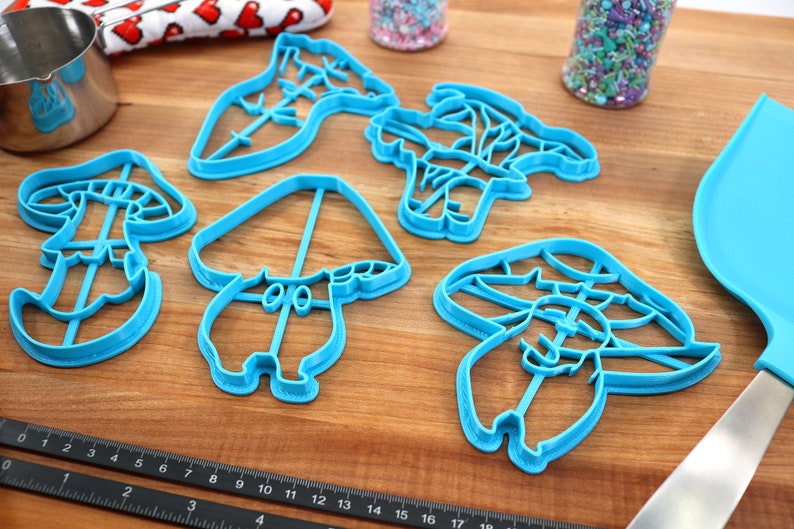 May include: Set of five blue plastic cookie cutters shaped like mushrooms. The cutters are on a wooden surface.