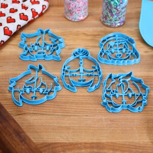 Puede incluir: Cinco cortadores de galletas de pl&aacute;stico azul con forma de caras de dibujos animados. Los cortadores est&aacute;n sobre una superficie de madera con un rodillo, un guante de horno rojo y blanco y una esp&aacute;tula azul.