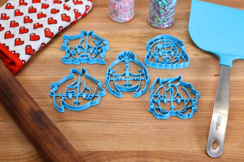 Puede incluir: Cinco cortadores de galletas de pl&aacute;stico azul con forma de caras de dibujos animados. Los cortadores est&aacute;n sobre una superficie de madera con un rodillo, un guante de horno rojo y blanco y una esp&aacute;tula azul.