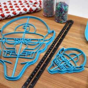 May include: Two blue plastic cookie cutters shaped like a cartoon character with a hat. The larger cutter is 15 inches wide and the smaller cutter is 6 inches wide. The cutters are on a wooden cutting board with a black ruler and a blue spatula.