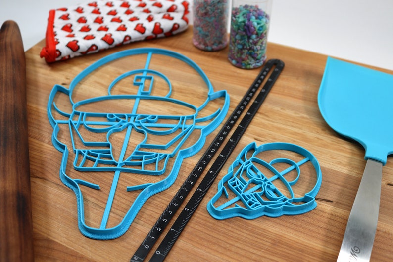 May include: Two blue plastic cookie cutters shaped like a cartoon character with a hat. The larger cutter is 15 inches wide and the smaller cutter is 6 inches wide. The cutters are on a wooden cutting board with a black ruler and a blue spatula.