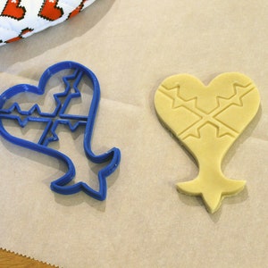 May include: A blue plastic cookie cutter shaped like a heart with a cross pattern inside, and a yellow cookie dough cutout in the same shape.