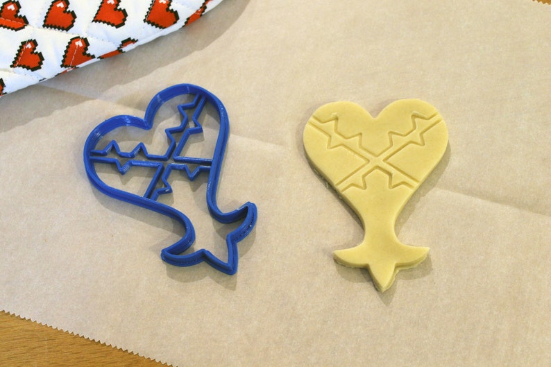 May include: A blue plastic cookie cutter shaped like a heart with a cross pattern inside, and a yellow cookie dough cutout in the same shape.