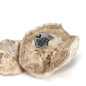 May include: A small, silver-colored metal pin with a shield-like design and a stylized emblem. The pin is displayed on a light-colored, textured stone against a white background. The pin's design features a series of vertical lines within a curved shape.