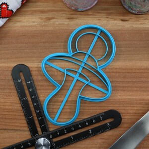 May include: A blue plastic cookie cutter shaped like a baby pacifier. The cutter is on a wooden surface with a ruler and a spatula.