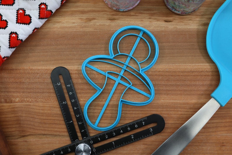 May include: A blue plastic cookie cutter shaped like a baby pacifier. The cutter is on a wooden surface with a ruler and a spatula.
