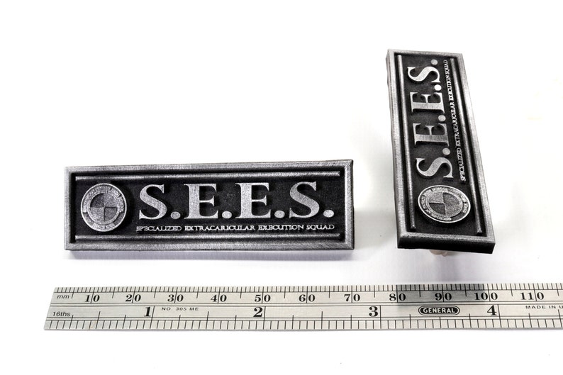 May include: Two silver metal badges with the text "S.E.E.S." and the words "SPECIALIZED EXTRACAJRICULAR DUBCUTION SQLLAD" on them. The badges are rectangular and have a raised border.