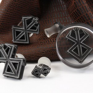 May include: Four metal stamps with a geometric design, resembling a stylized letter X, are displayed on a brown leather surface. The stamps are attached to clear plastic handles.