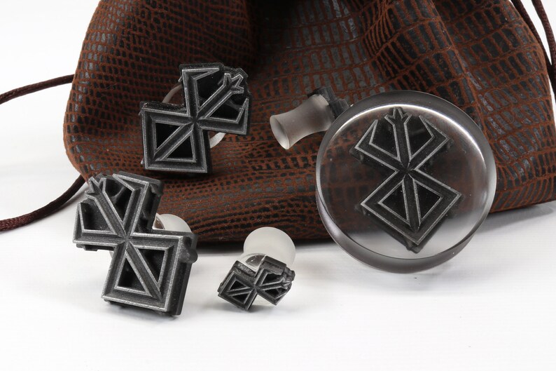 May include: Four metal stamps with a geometric design, resembling a stylized letter X, are displayed on a brown leather surface. The stamps are attached to clear plastic handles.