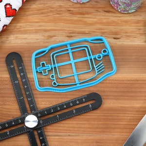 May include: A blue plastic cookie cutter shaped like a handheld video game console. The cutter has a detailed design with buttons and a screen.