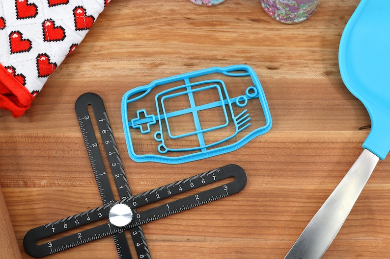 May include: A blue plastic cookie cutter shaped like a handheld video game console. The cutter has a detailed design with buttons and a screen.