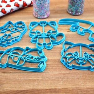 May include: Five blue plastic cookie cutters shaped like red pandas. The cutters are on a wooden surface.