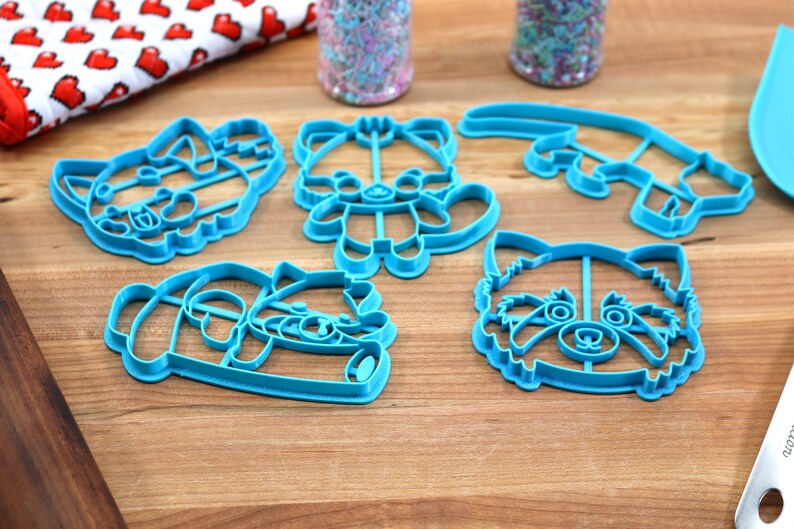May include: Five blue plastic cookie cutters shaped like red pandas. The cutters are on a wooden surface.