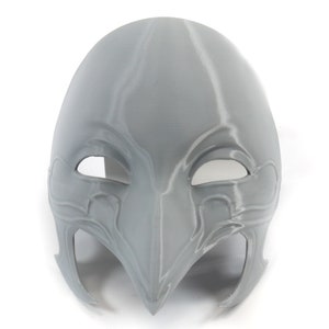 May include: A gray 3D printed mask with intricate details and eye holes. The mask has a pointed beak and a design that resembles a bird's head.