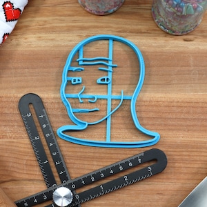 Op de afbeelding: Een blauwe plastic koekjesvormer in de vorm van de "Drakeposting" meme, een populaire internetmeme met het gezicht van een persoon met een gespleten uitdrukking. De koekjesvormer ligt op een houten oppervlak.
