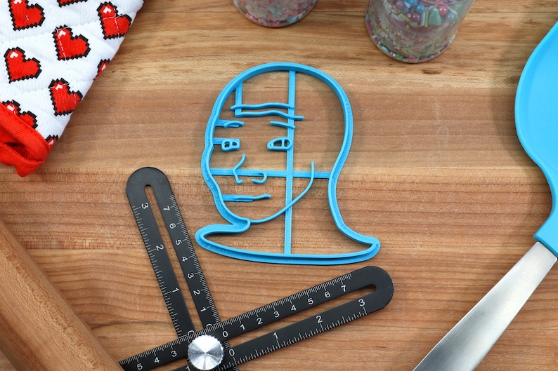 Op de afbeelding: Een blauwe plastic koekjesvormer in de vorm van de "Drakeposting" meme, een populaire internetmeme met het gezicht van een persoon met een gespleten uitdrukking. De koekjesvormer ligt op een houten oppervlak.