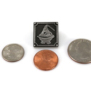 May include: A silver metal pin with a black background and a silver design of a stylized genie lamp. The pin is about the size of a US quarter.