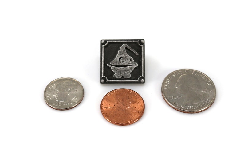 May include: A silver metal pin with a black background and a silver design of a stylized genie lamp. The pin is about the size of a US quarter.