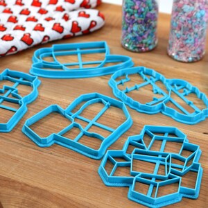 May include: A set of six blue plastic cookie cutters shaped like a teapot, mug, cup and saucer, a snowflake, a house, and a gift box. The cutters are on a wooden surface.